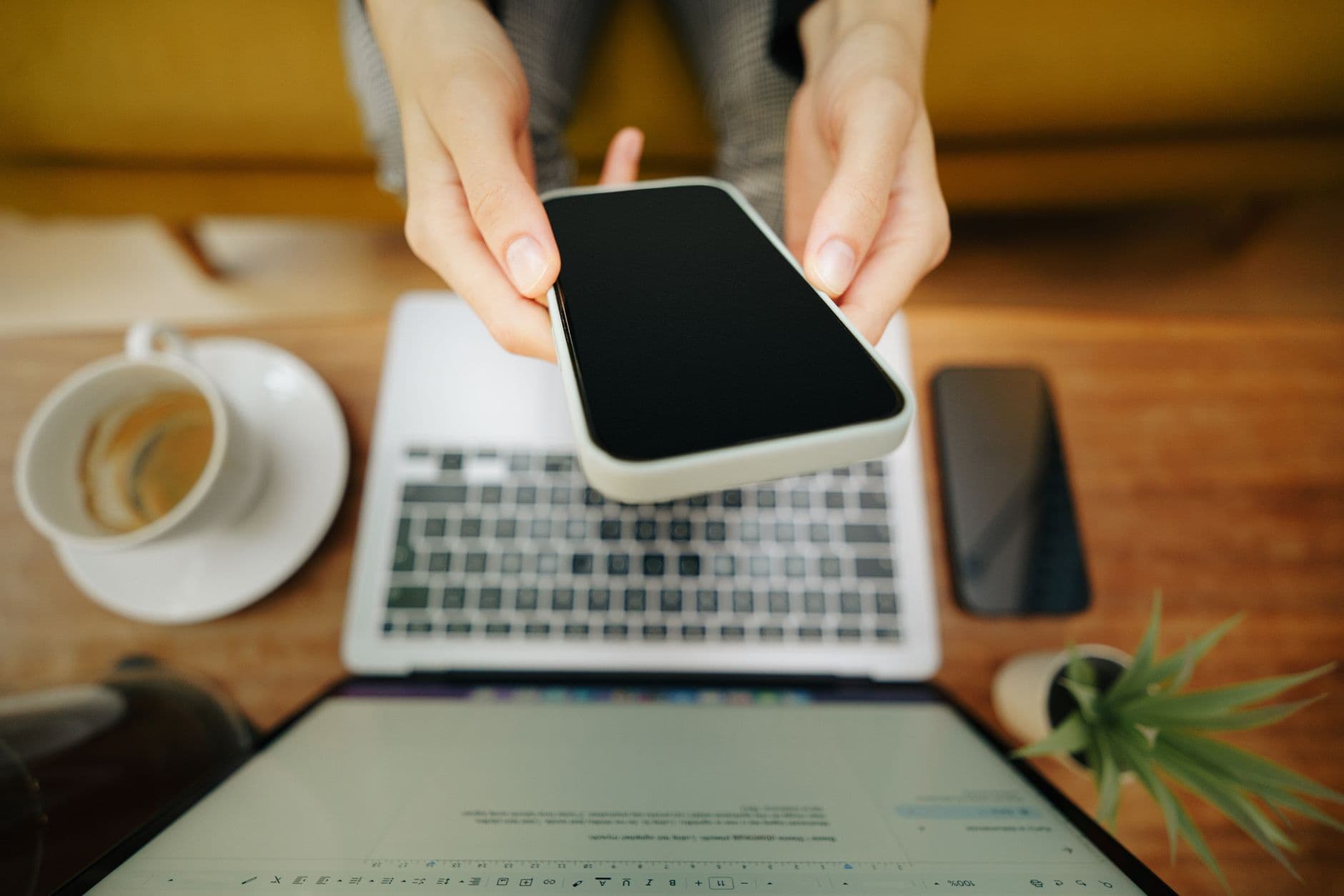 A pair of hands holding a smartphone with a laptop on the table